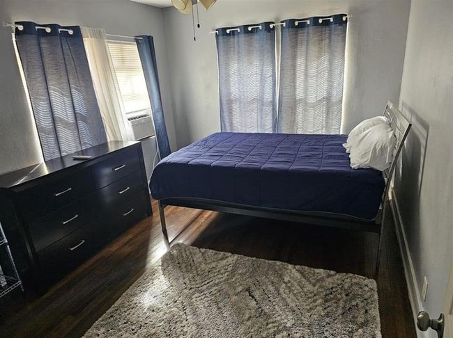 This cozy bedroom features hardwood floors, a comfortable bed, and ample natural light. It includes a dark wood dresser and a soft area rug, with blue curtains complementing the neutral wall color. | Image 25