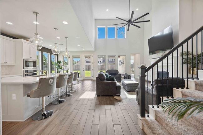 This modern living space features an open floor plan with a spacious kitchen, complete with a large island and elegant pendant lighting. The living area boasts high ceilings, large windows for ample natural light, and a contemporary ceiling fan. A sleek staircase leads to the upper level, adding to the home's sophisticated design. | Image 9
