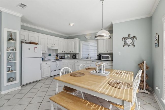 This bright and inviting eat-in kitchen features crisp white cabinetry, ample counter space, and a convenient layout perfect for both meal prep and casual dining. A large window over the sink lets in natural light, enhancing the airy feel of the space. The adjoining dining area comfortably accommodates a spacious table, making it ideal for family meals or entertaining. Tile flooring throughout adds durability and ease of maintenance, while the soft coastal color palette ties the kitchen seamlessly into the | Image 6