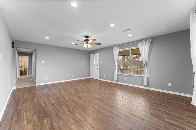 Bright and spacious living room with wood-look tile flooring, neutral gray walls, recessed lighting, and a ceiling fan. Large window with sheer curtains offers natural light and a view of the outdoors. | Image 11