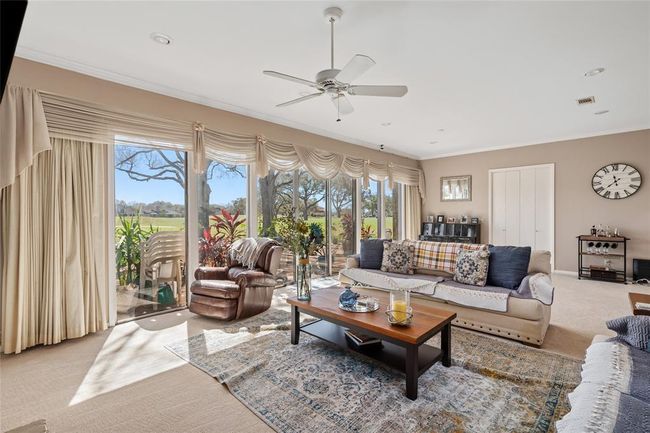 This spacious living room offers a warm and inviting atmosphere, featuring large windows that frame the backyard pergola and patio area. Plush carpeting, soft neutral tones, and elegant crown molding enhance the room’s charm, while the open layout provides flexibility for entertaining or relaxing. | Image 12