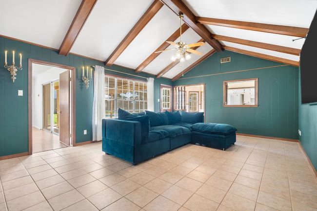 Living area with wood panel walls, ceiling fan, and light tile patterned flooring. The windows overlook the covered backyard patio and pool. The door to the left opens up to the primary bedroom. | Image 15