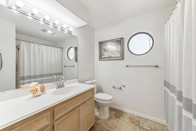 Bright bathroom with a large mirror, modern lighting, and circular window. Features a shower-tub combo with a curtain, tiled floor, and wooden vanity with a single sink. | Image 21