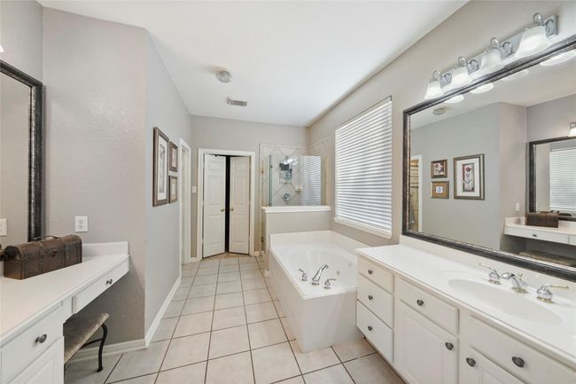 This spacious bathroom features a large soaking tub, dual sinks with ample counter space, and a makeup vanity. The room is brightened by a large window, and the neutral tiles and walls give it a fresh, modern feel. | Image 20