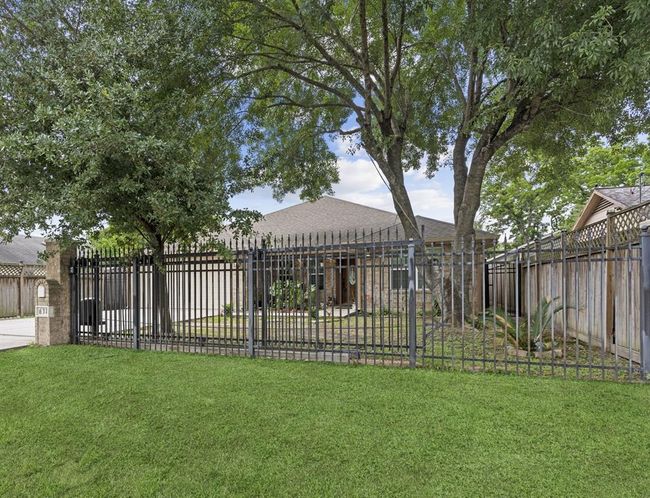 The photo shows a well-maintained single-story home with a sizable, lush lawn. Mature trees provide shade and privacy. The property is secured with an ornamental iron fence, enhancing both security and curb appeal. | Image 4