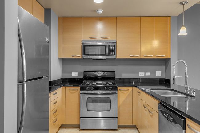 Kitchen featuring light brown cabinetry, dark stone countertops, a sink, decorative light fixtures, and appliances with stainless steel finishes | Image 6