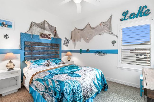This bedroom features a nautical theme with ocean-inspired decor, including fishing nets and maritime accents. It has a cozy bed with blue and white bedding, a wooden headboard, and coastal decorations. The room is bright with natural light coming through the window shutters, creating a serene, beachy atmosphere with 2 new picture windows and view of the lake. | Image 24