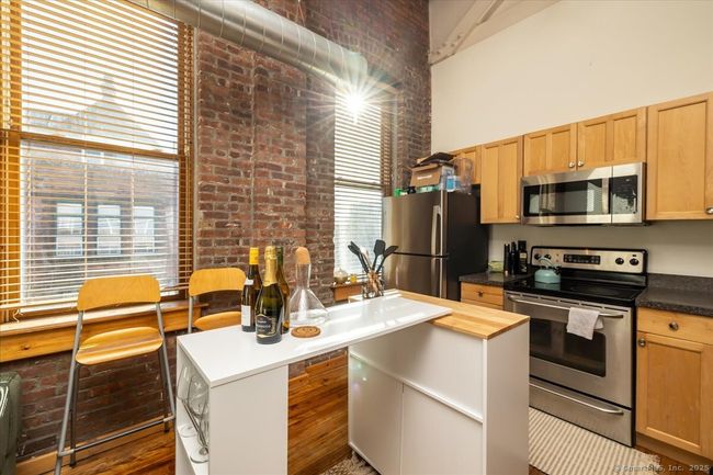 Modern-industrial kitchen with exposed brick walls, high ceilings, and large windows allowing ample natural light, featuring wooden cabinetry and stainless steel appliances | Image 7