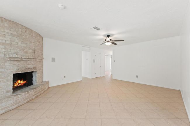 This spacious living area features a cozy brick fireplace, neutral tile flooring, and a ceiling fan, offering a comfortable and inviting atmosphere. | Image 10