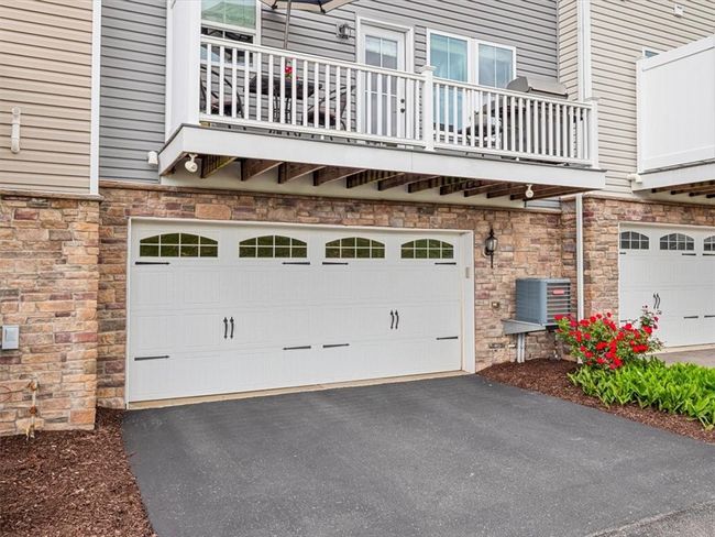 This townhome features a spacious two-car garage with charming carriage-style doors and decorative windows, offering both functionality and curb appeal. The brick façade adds a touch of elegance and durability, while the overhead balcony provides outdoor living space directly above. A freshly paved driveway and neatly landscaped garden bed complete the exterior, making for a well-maintained and welcoming entrance to the home. | Image 22