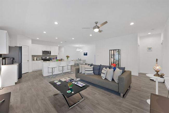 This photo showcases a modern open-concept living space with a sleek kitchen featuring white cabinetry, stainless steel appliances, and a spacious island with bar seating. The adjoining living area is bright, furnished with a comfortable sofa and stylish decor, accented by wood-look flooring and recessed lighting. | Image 11
