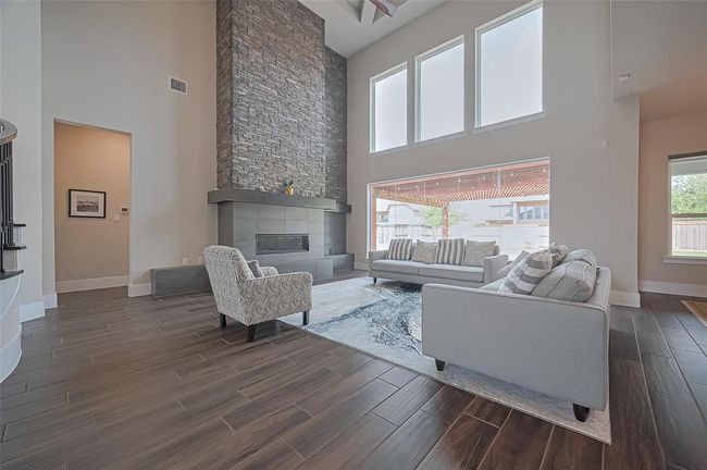 This spacious living room features a dramatic floor-to-ceiling stone fireplace, high ceilings, and large windows that fill the space with natural light. The open layout is accented by elegant wood tile flooring and stylish furnishings, offering a modern and inviting atmosphere. | Image 8