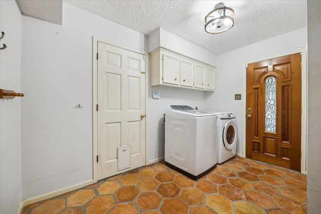 First floor laundry room with overhead cabinets for storage, and a charming wooden door with decorative glass. The space has saltillo tile flooring and a modern ceiling light fixture. Two doors, one going outside and the other going into the garage. | Image 27