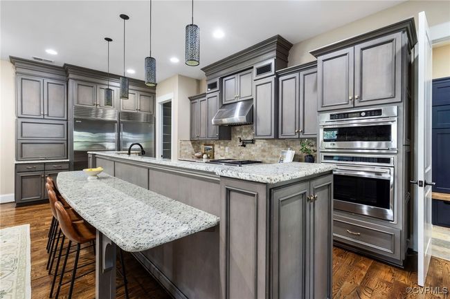 Kitchen featuring a spacious island, dark wood-style flooring, under cabinet range hood, backsplash, and light stone countertops | Image 9