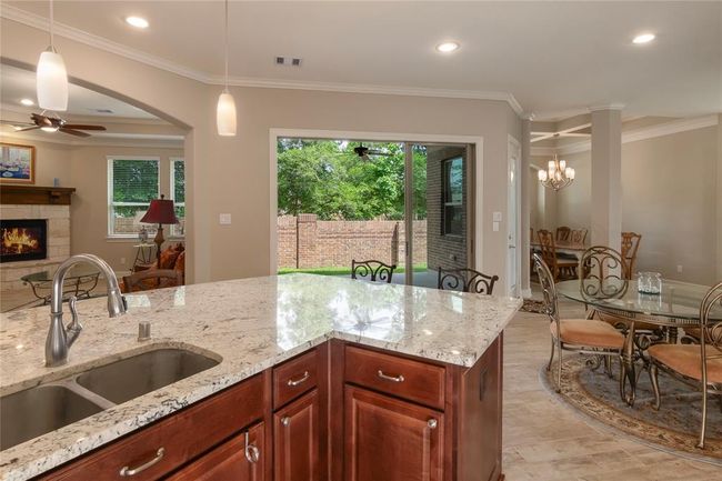 This photo showcases a spacious open-concept kitchen and living area. It features a large granite island with a double sink, rich wood cabinetry, and recessed lighting. The space flows into a cozy living room with a fireplace and a dining area with a stylish chandelier. Large windows and a glass door provide natural light and access to a fenced backyard, offering a seamless indoor-outdoor experience. | Image 14