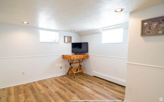 Basement featuring a baseboard heating unit, a textured ceiling, wood finished floors, wainscoting, and healthy amount of natural light | Image 13