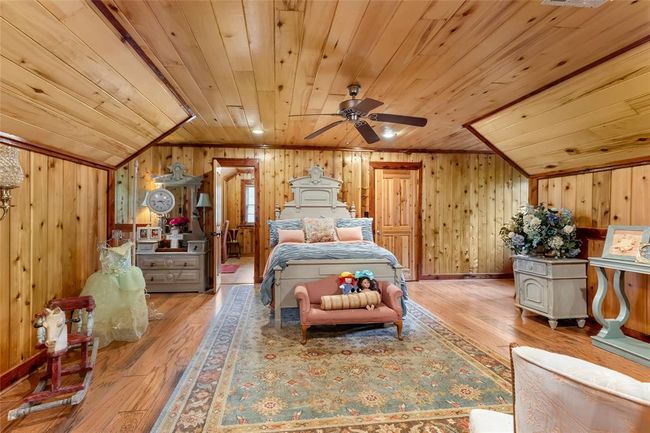 This cozy bedroom features charming wood-paneled walls and ceiling, creating a warm, rustic feel. It includes a central bed, classic furniture, and a decorative area rug, complemented by a ceiling fan for comfort. | Image 40