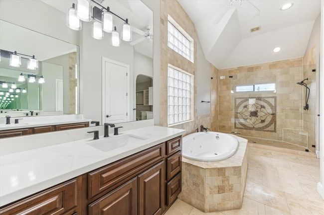 This bathroom features a spacious layout with a luxurious whirlpool tub, large walk-in shower with intricate tile design, and dual sinks with modern fixtures. The room is accented with ample natural light from glass block windows and sleek lighting fixtures. | Image 5