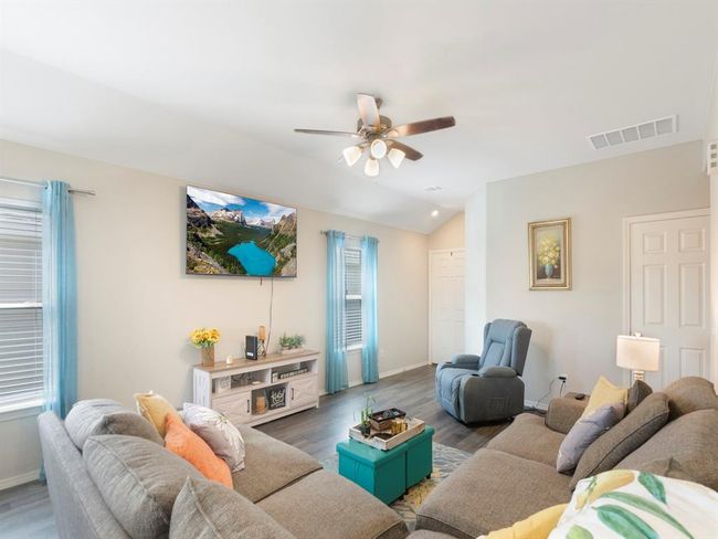 Bright and inviting living room with a cozy sectional sofa, a recliner, and a wall-mounted TV. The space features vinyl plank floors, light walls, and abundant natural light from two windows with sheer curtains. The room is tastefully decorated with a modern ceiling fan, artwork, and a small entertainment unit. | Image 4