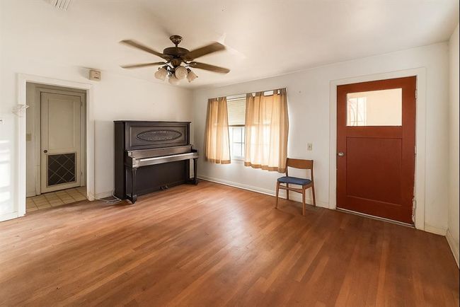 Bright room featuring hardwood floors and a ceiling fan, with a window letting in natural light. Includes a piano and a chair, offering a cozy atmosphere. Access through a wooden door and an adjacent hallway. | Image 4