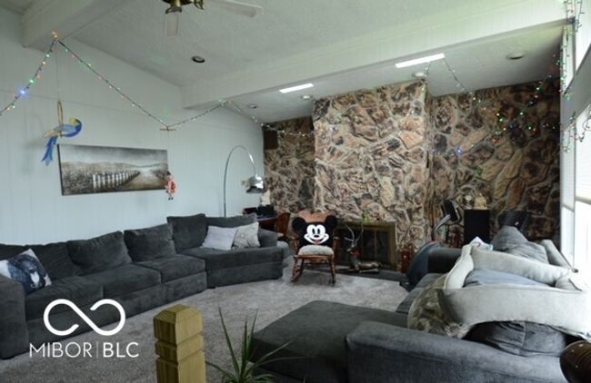 carpeted living room with beamed ceiling, a ceiling fan, and a stone fireplace | Image 9
