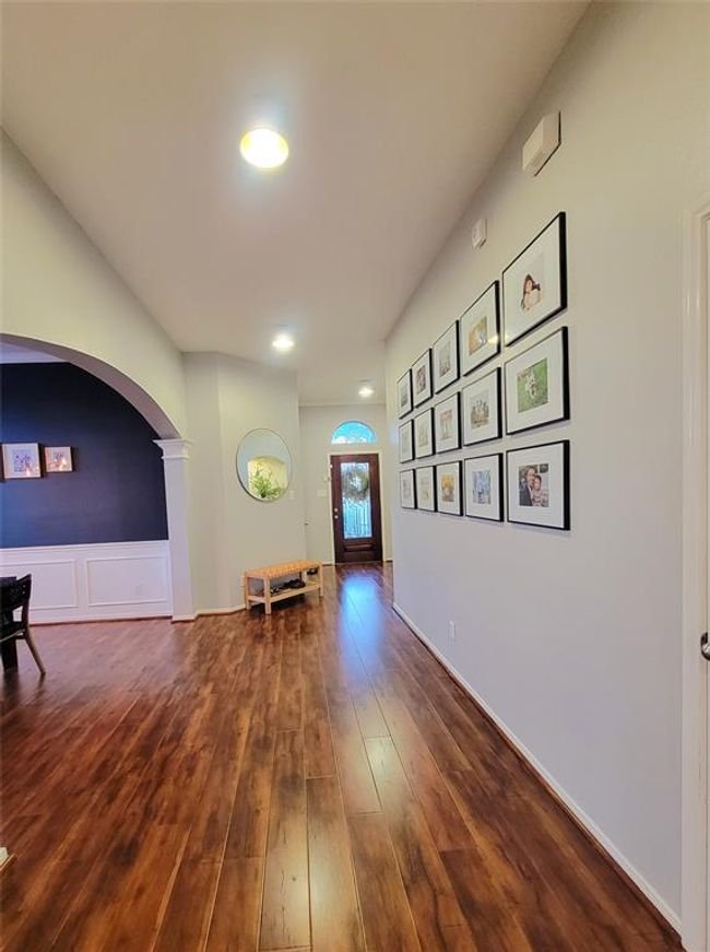Bright and welcoming entryway with wood flooring, recessed lighting, and a gallery wall. The archway leads to a dining area with wainscoting and a dark accent wall. A glass-paneled front door adds elegance. | Image 16