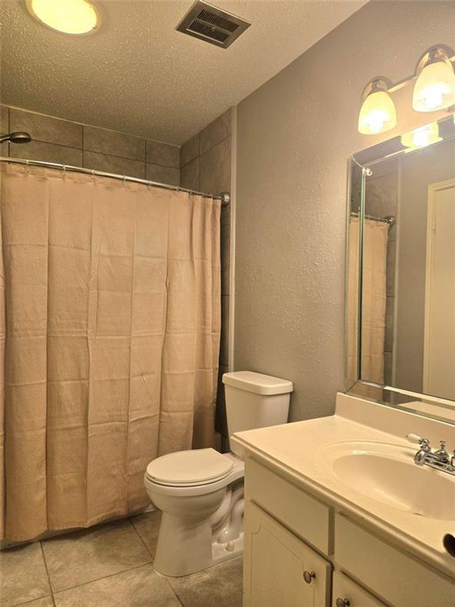 Experience a cozy and inviting bathroom featuring a tiled shower, beige curtain, and a sleek vanity. Its neutral tones and soft lighting create a welcoming ambiance. | Image 12