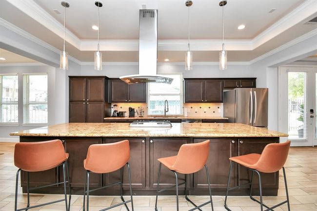 This kitchen boasts a spacious open layout with a large central island featuring granite countertops and stylish pendant lighting. It includes modern stainless steel appliances, ample dark wood cabinetry, and seating for four. Natural light floods in through large windows, creating a bright and inviting space. | Image 10