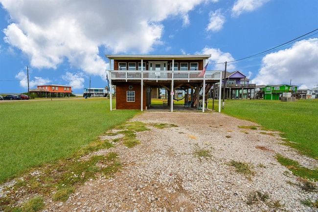 This photo shows a raised beach house with a spacious front porch, set on a grassy lot. The house has a gravel driveway and is elevated on stilts, typical for coastal areas. It's surrounded by other colorful elevated homes, with a bright, open sky above. | Image 33