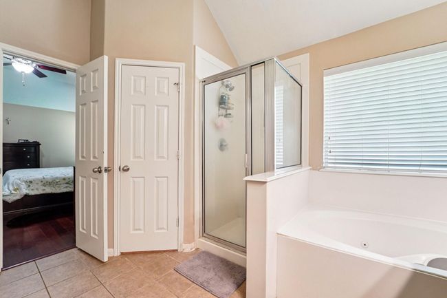 This bathroom features a glass-enclosed shower, a separate bathtub, and neutral tiles. It connects to a bedroom with wooden flooring. Natural light filters through a window with blinds, creating a bright and inviting space. | Image 16