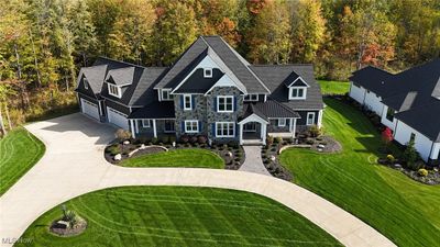 An aerial shot offering a broad perspective of the property, including the main house, driveway, and surrounding greenery. The home sits on a spacious, well-maintained lot bordered by a wooded area, with the driveway curving gracefully to the attached four-car garage. | Image 1