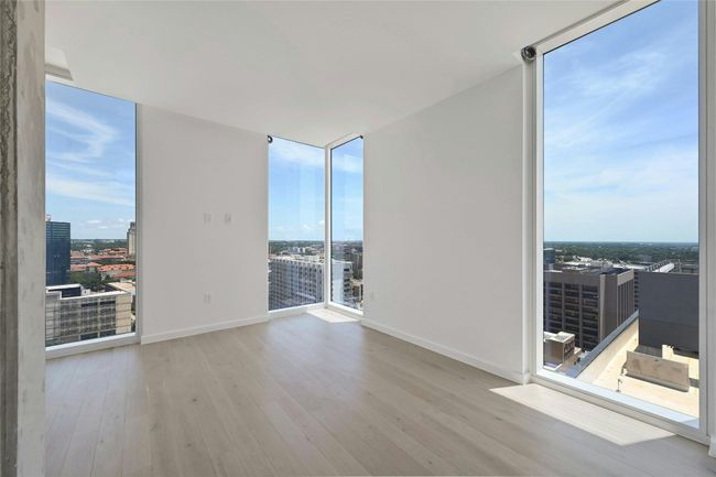 Stunning primary bedroom featuring dramatic floor-to-ceiling corner windows that flood the space with natural light and showcase panoramic downtown views. Light-toned hardwood floors and clean modern finishes enhance the bright, minimalist aesthetic—perfect for a serene city retreat. | Image 7