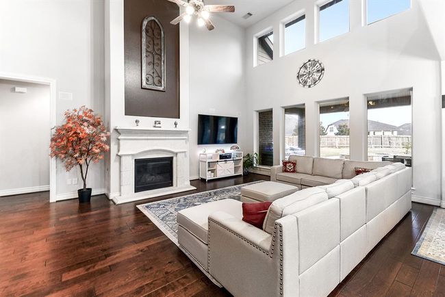 This expansive living room features high ceilings and eight large windows that flood the space with natural light. The same beautiful wood flooring found throughout the rest of the home extends into this room, adding warmth and continuity. The gas cast stone fireplace serves as a striking focal point, creating a cozy and inviting atmosphere. | Image 18