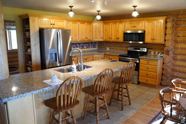 Kitchen with appliances with stainless steel finishes, log walls, a breakfast bar, decorative light fixtures, and light stone counters | Image 10