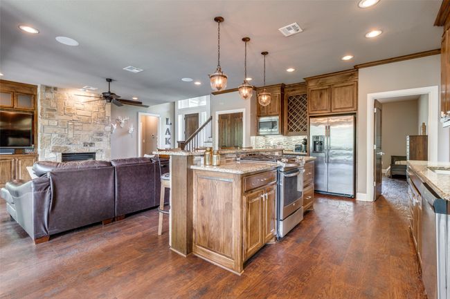 Kitchen with light stone countertops, a stone fireplace, stainless steel appliances, dark wood-type flooring, and a kitchen island with sink | Image 7
