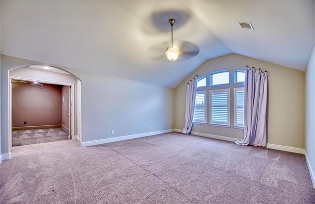This spacious room features a vaulted ceiling with a ceiling fan, neutral carpeting, and a large arched window with curtains, offering plenty of natural light. An open doorway leads to an adjacent room, enhancing the sense of space. | Image 43