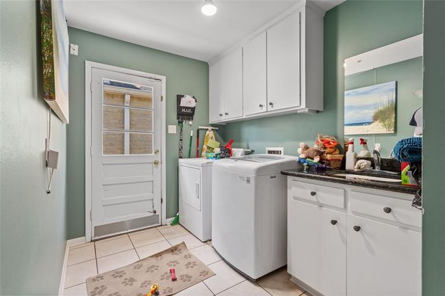 This functional laundry room offers ample storage and workspace with upper and lower white cabinetry, a built-in sink, and granite countertops. The washer and dryer are conveniently positioned, while the door provides easy access to the backyard. The space is accented by green walls, tiled flooring, and a practical layout designed for efficiency. | Image 22