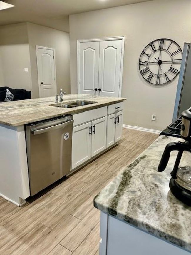 Discover a modern kitchen with granite countertops, a spacious island with a sink, stainless steel appliances, and elegant wood-look tile flooring. How would you decorate the wall with the stylish wall clock. | Image 8