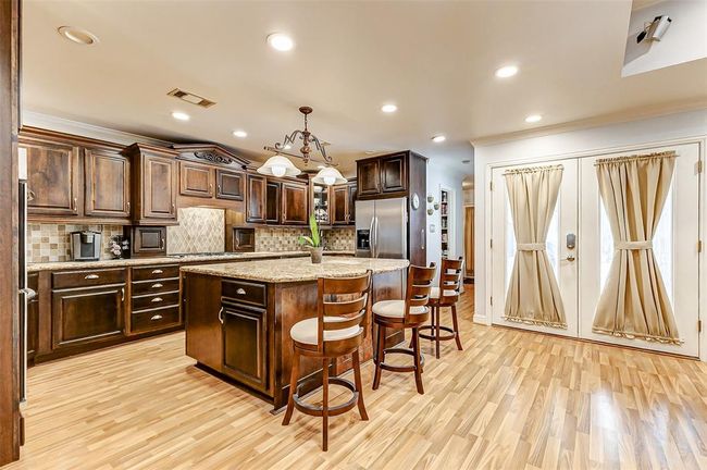 A chef’s dream, this kitchen seamlessly blends elegance with functionality. Rich dark wood cabinetry with ornate accents offers ample storage, complemented by extensive countertops and an island with seating. High-end features include two sinks, two dishwashers, a wine fridge, stainless steel appliances, and a custom pantry designed for ultimate convenience. A stylish backsplash and elegant pendant lighting complete the space. | Image 12
