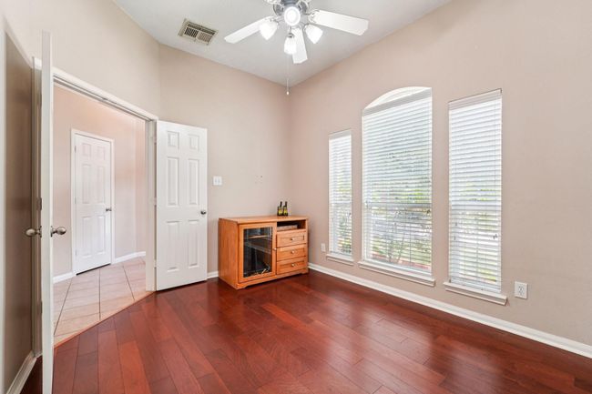 Bright room with hardwood floors, high ceiling, and large windows for natural light. Features a ceiling fan and connects to a tiled hallway. | Image 6