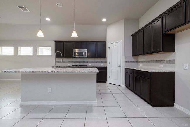 Kitchen featuring stainless steel microwave, backsplash, light stone counters, light tile patterned flooring, and a center island with sink | Image 7