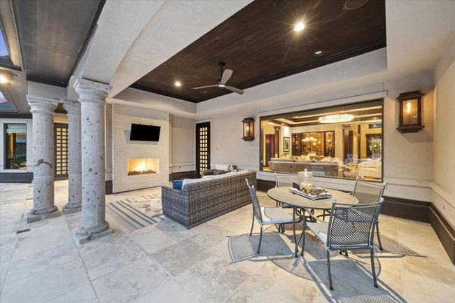 This elegant patio is the ultimate extension of indoor-outdoor living, complete with a gas fireplace with stacked stone surround, gas lanterns and mounted TV for year-round enjoyment. Framed by stately columns and highlighted by a rich wood ceiling with recessed lighting, built-in speakers and a ceiling fan, the space blends sophistication and comfort. Ideal for both lounging and al fresco dining, it’s a seamless connection to the interior entertaining areas just beyond the oversized window. | Image 45