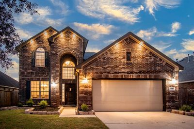 This is a beautiful two-story brick residence featuring an arched entryway, tall windows, and a two-car garage, captured at dusk with custom exterior lighting accentuating its architectural details. | Image 1