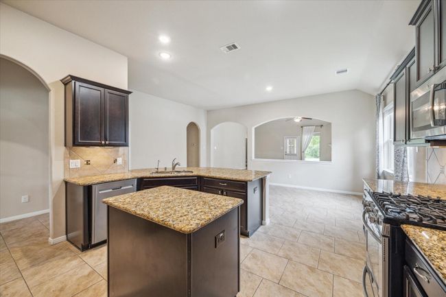 This kitchen features dark wood cabinetry, granite countertops, a central island, and stainless steel appliances. It offers a spacious layout with tiled flooring and recessed lighting, perfect for modern living. | Image 15
