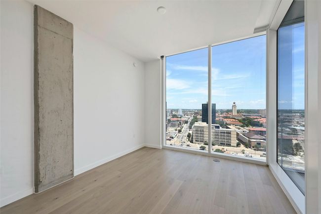 Step into this sun-drenched bedroom featuring expansive floor-to-ceiling windows that offer breathtaking panoramic views of the city skyline. The clean, modern design is highlighted by light hardwood flooring, crisp white walls, and a striking exposed concrete column that adds a touch of industrial elegance. | Image 11