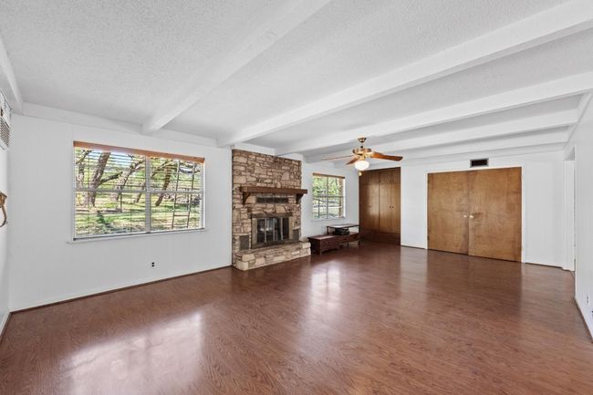 Unfurnished living room featuring a stone fireplace, beam ceiling, wood finished floors, and a ceiling fan | Image 7