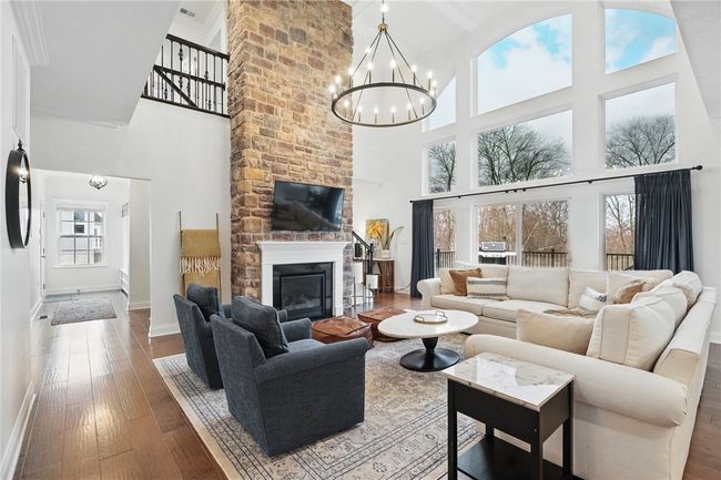 Stunning two-story great room featuring a floor-to-ceiling stone fireplace, dramatic wall of windows with woodland views, and a grand chandelier. Open and airy layout with hardwood floors and abundant natural light, creating a warm and inviting space ideal for relaxing or entertaining. | Image 9