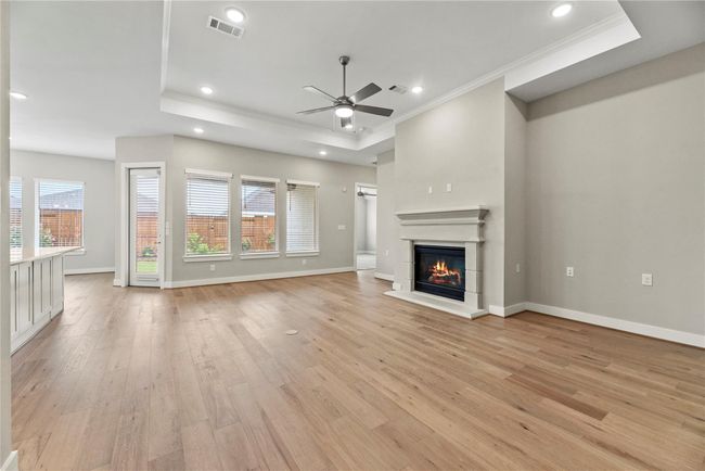 This inviting family room features warm LVP flooring, a cozy gas fireplace, and a stylish tray ceiling with recessed lighting and a modern fan. Natural light pours in through a wall of windows, creating a bright and open space that’s perfect for everyday living and relaxed entertaining. | Image 9