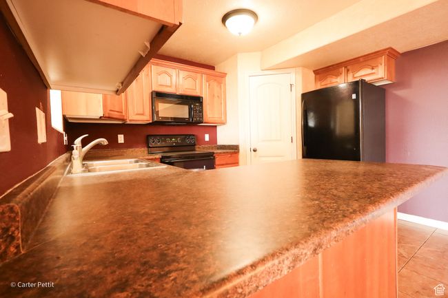 Kitchen with black appliances, light brown cabinetry, light tile patterned floors, a peninsula, and dark countertops | Image 22