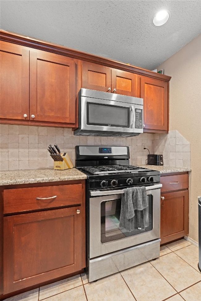 Kitchen featuring appliances with stainless steel finishes, a textured ceiling, tasteful backsplash, brown cabinetry, and light stone countertops | Image 6